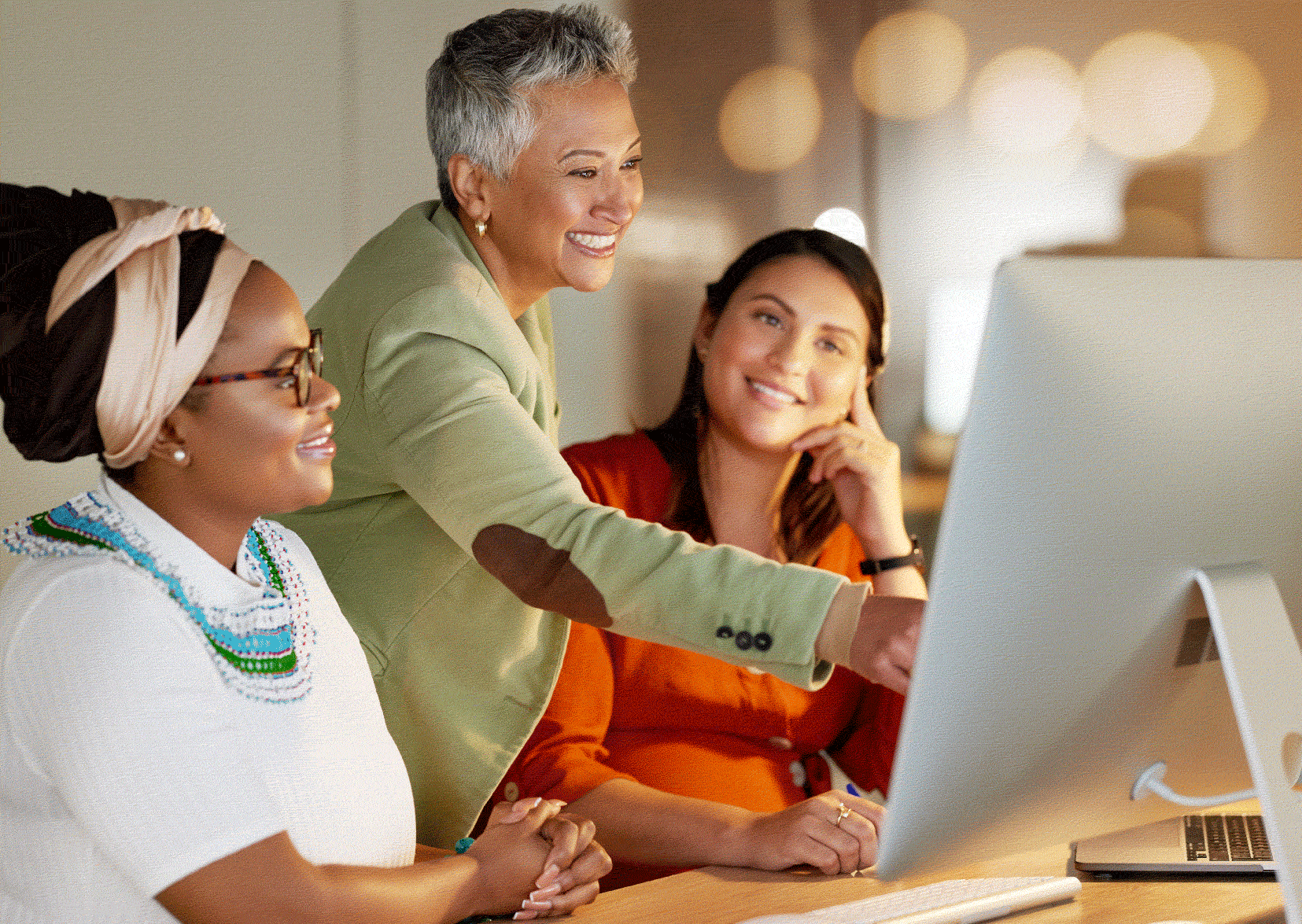 Three women smiling looking at a Mac screen. One pointing her finger